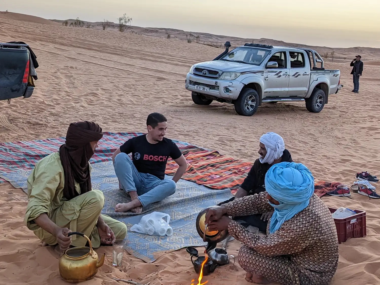 Travellers sitting around a fire on a rug in the desert at dawn near El Menia
